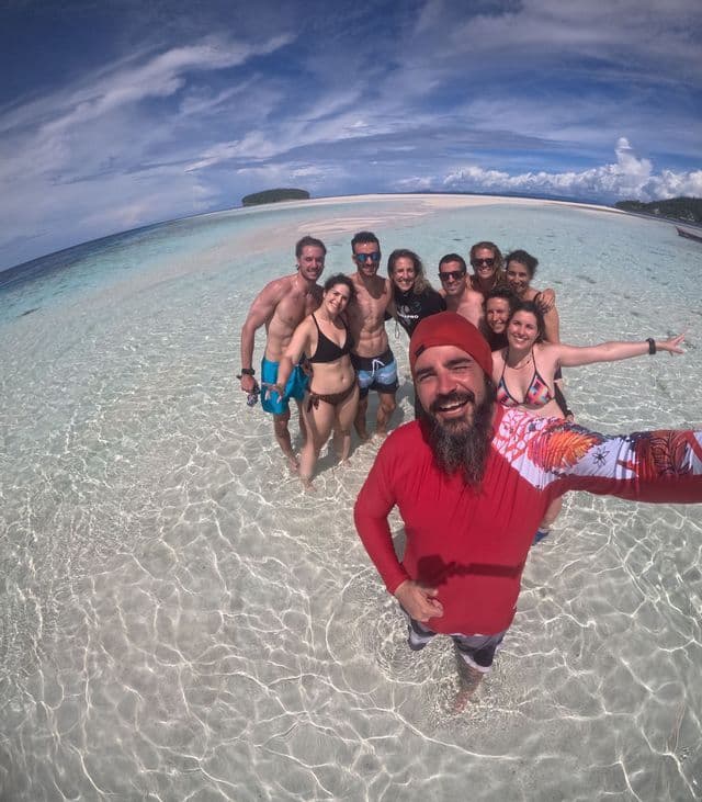 Un hombre sonriente de camiseta roja se hace una selfie con un grupo de WeRoad en aguas claras y poco profundas del mar.