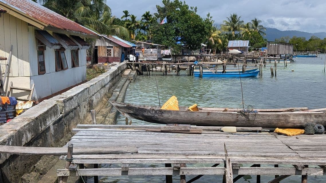 Una canoa de madera amarrada a un muelle en un pueblo costero con casas sobre pilotes y colinas al fondo.