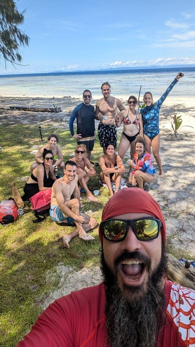 Un hombre barbudo se toma un selfie sonriente con un grupo de WeRoad posando en una playa tropical y soleada junto al mar.