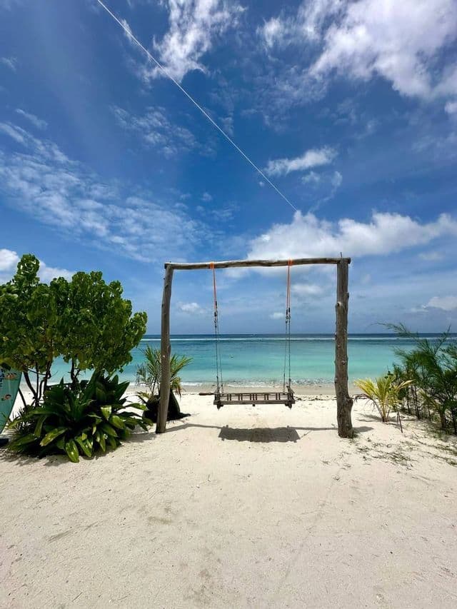 Un'altalena di legno si trova su una spiaggia di sabbia bianca, di fronte a un oceano turchese sotto un cielo blu con nuvole sparse.