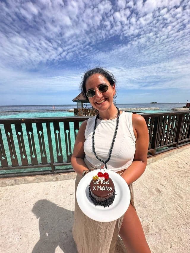 Una donna sorridente con occhiali da sole tiene in mano una torta al cioccolato su un piatto con la scritta 'We Road X Maldive', mentre si trova su una spiaggia con acqua turchese.
