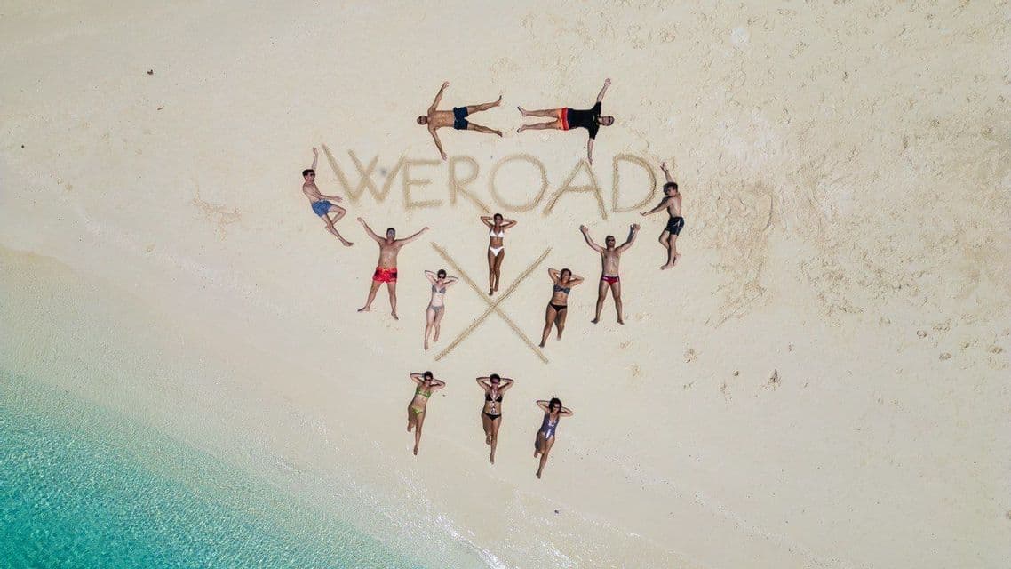 Veduta aerea di un gruppo WeRoad sdraiato su una spiaggia di sabbia bianca attorno alla parola 'WEROAD' scritta sulla sabbia