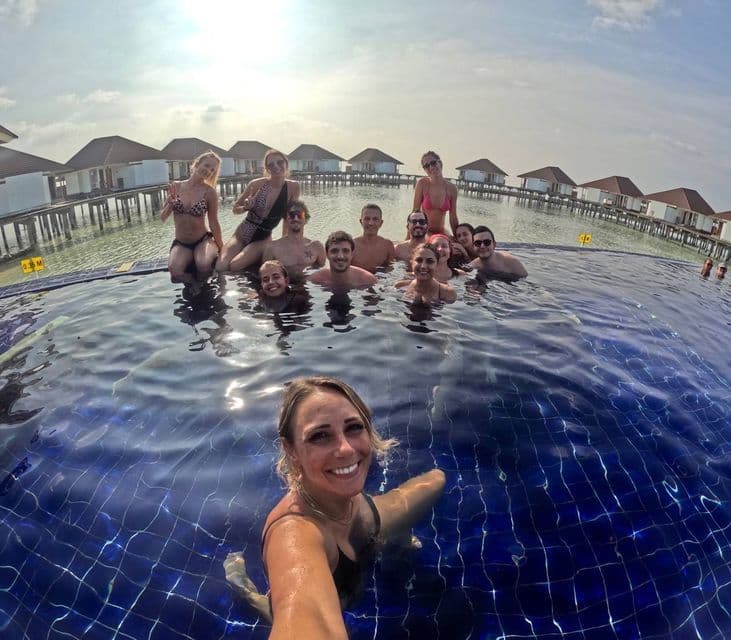 Una donna si scatta un selfie con un gruppo WeRoad, in una piscina a sfioro con bungalow sull'acqua sullo sfondo.