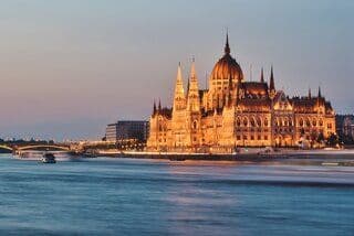 An ornate government building with a large dome and spires, illuminated with golden lights at dusk on a riverbank.