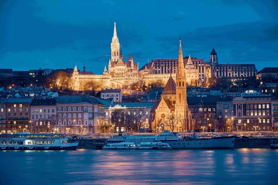 A historic European city skyline with illuminated churches and buildings seen from across a river at dusk.