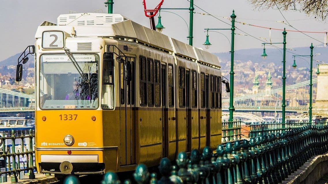 Eine gelb-weiße Straßenbahn der Linie 2 fährt auf Gleisen in einer Stadt, im Hintergrund sind eine Brücke und Gebäude zu sehen.