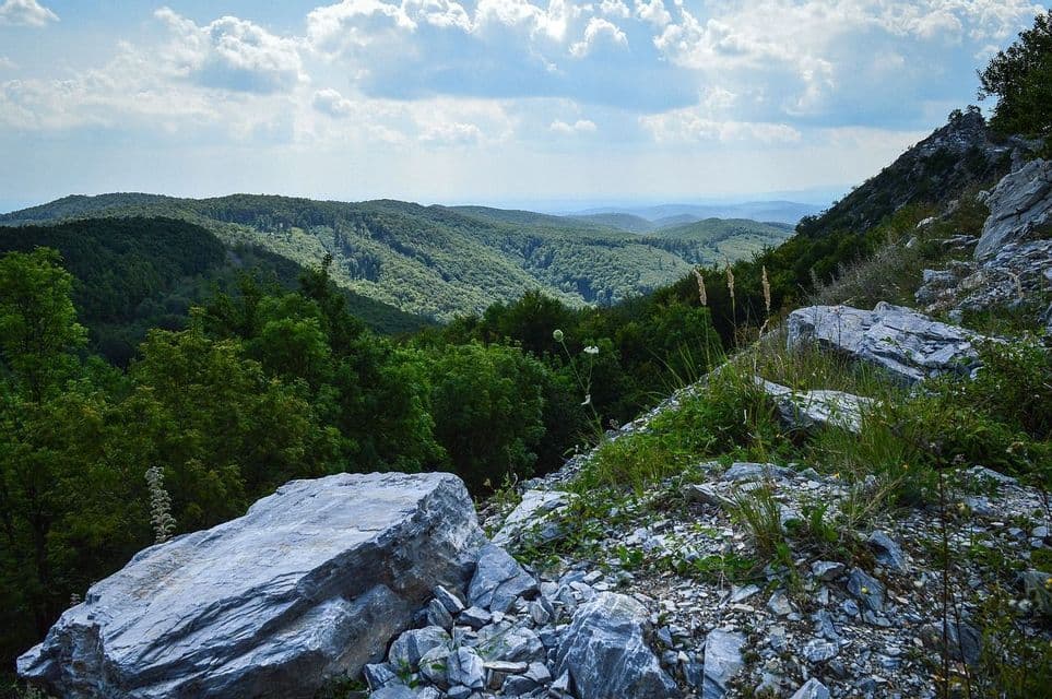 Ein Blick von einem felsigen Berghang auf sanfte, von grünem Wald bedeckte Hügel unter einem blauen Himmel mit weißen Wolken.