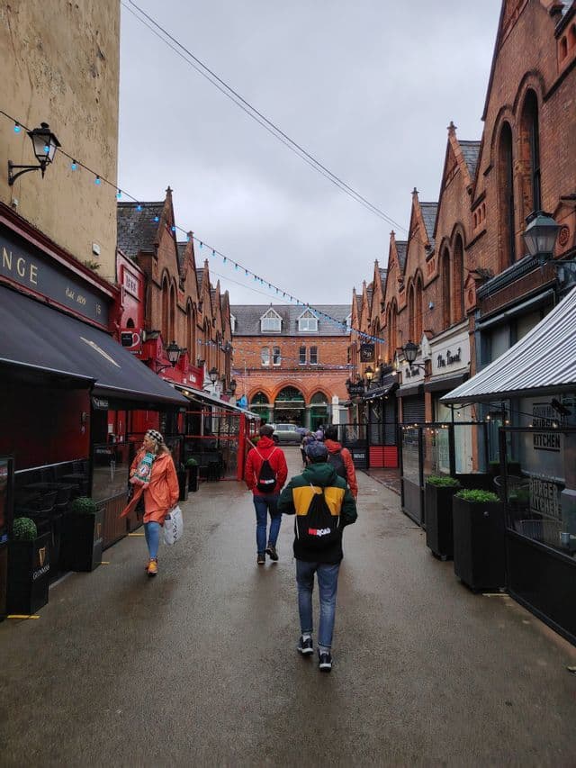 A WeRoad group trip walks down a wet, narrow street lined with red brick shops and pubs under an overcast sky.