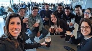 A WeRoad group trip smiles for a selfie while holding glasses of beer around a table indoors.