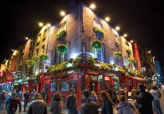 A brightly lit red pub on a brick building corner at night, with a crowd of people walking on the street below.