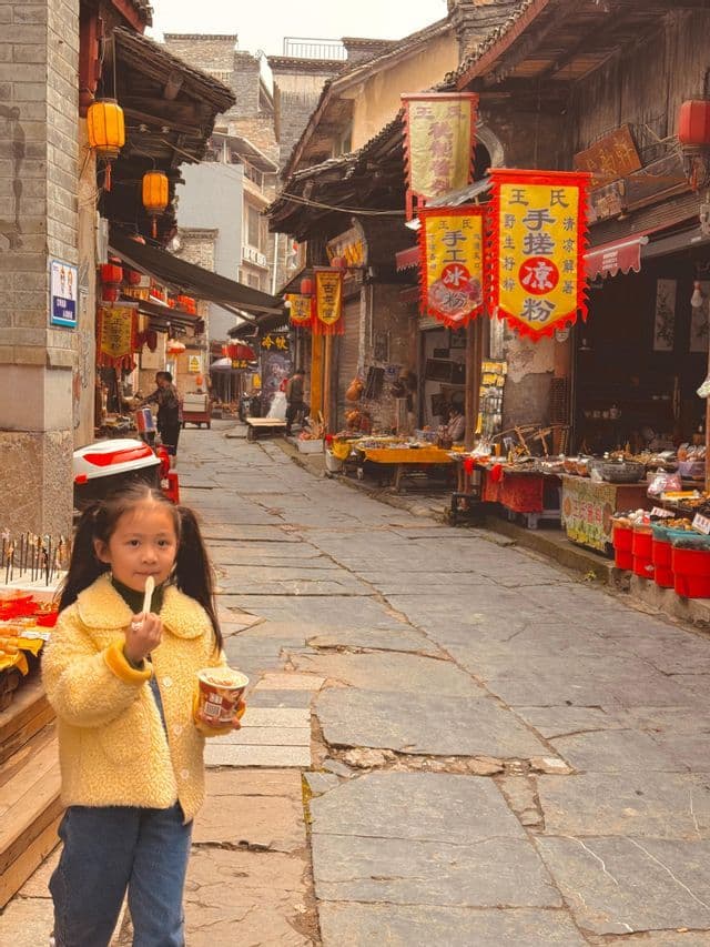 Una giovane ragazza con una giacca gialla mangia da una tazza su una stretta via acciottolata in un mercato.