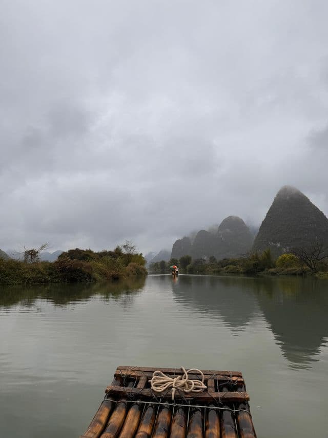 Vista dalla parte anteriore di una zattera di bambù su un fiume calmo, con montagne carsiche avvolte nella nebbia sotto un cielo pesante e coperto.
