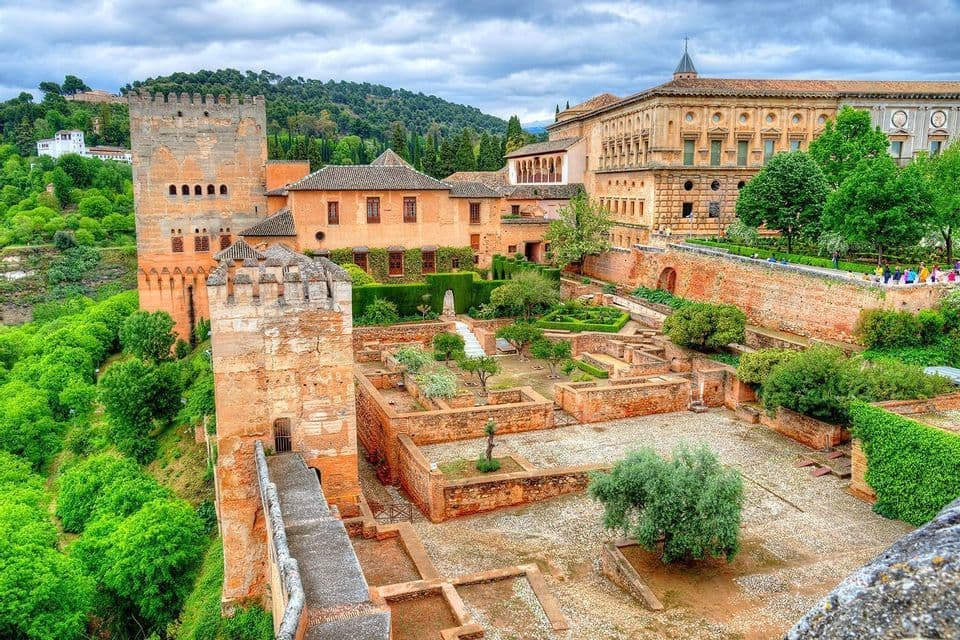 Una vista dall'alto di una fortezza storica con torri in pietra e giardini terrazzati, che domina una collina ricoperta di alberi lussureggianti.