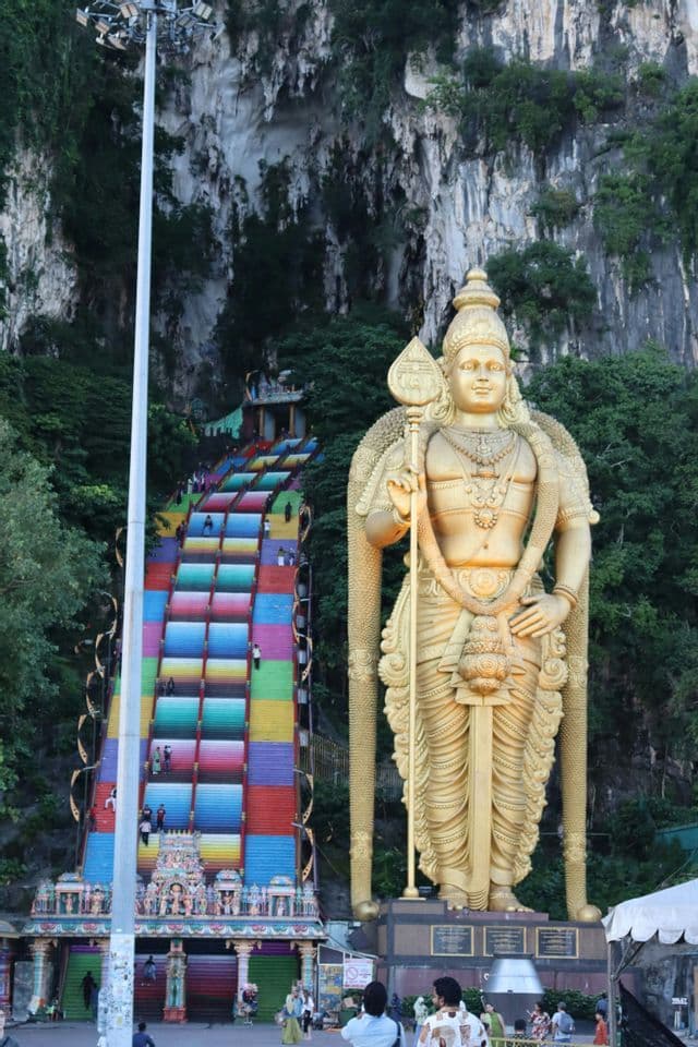 Eine riesige goldene Statue einer Gottheit steht neben einer langen, farbenfrohen Treppe, die eine felsige Klippe hinaufführt, mit Menschen auf den Stufen.