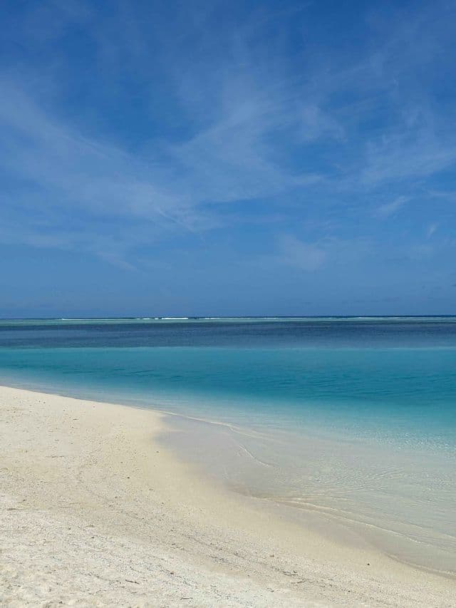 Una spiaggia di sabbia bianca incontra l'oceano limpido color turchese e blu profondo sotto un vasto cielo azzurro con nuvole leggere.