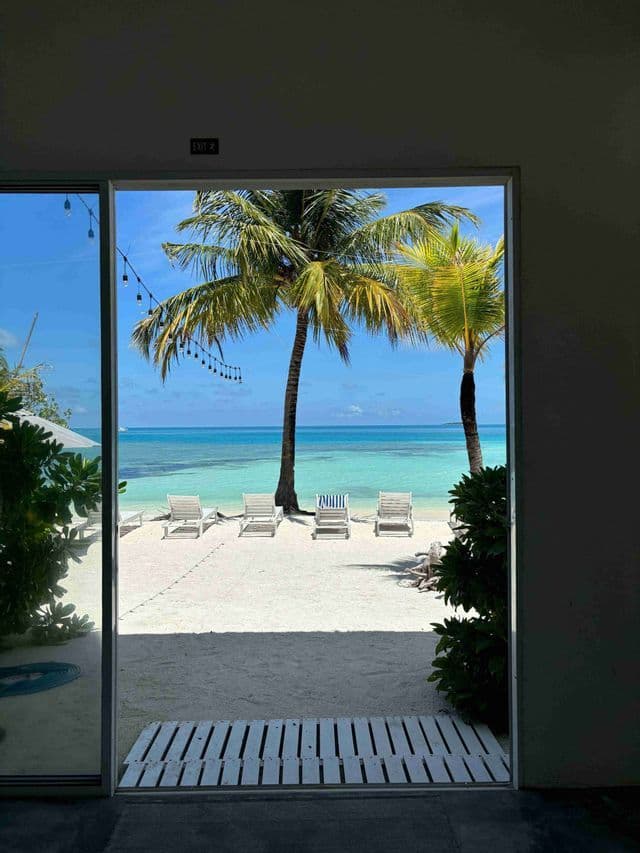 Vista da un'apertura su una spiaggia di sabbia bianca con palme e lettini che si affacciano sull'acqua turchese.