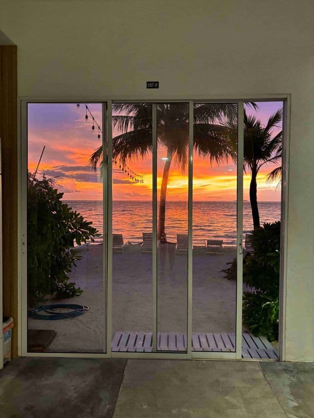 Un tramonto vibrante su una spiaggia tropicale con palme, visto dall'interno attraverso una grande porta scorrevole in vetro.