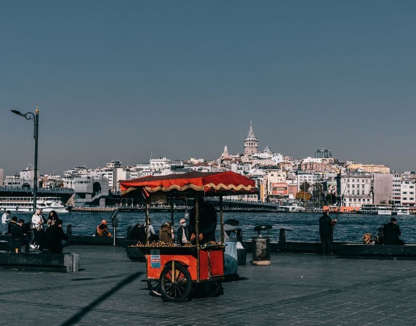 Un carrito rojo de comida callejera en un paseo marítimo con un paisaje urbano y una torre visibles al fondo al otro lado del agua.