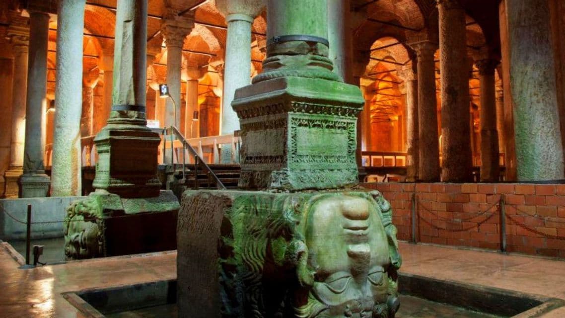 Una cabeza de piedra tallada invertida sirve de base de columna en una antigua cisterna subterránea llena de columnas iluminadas.