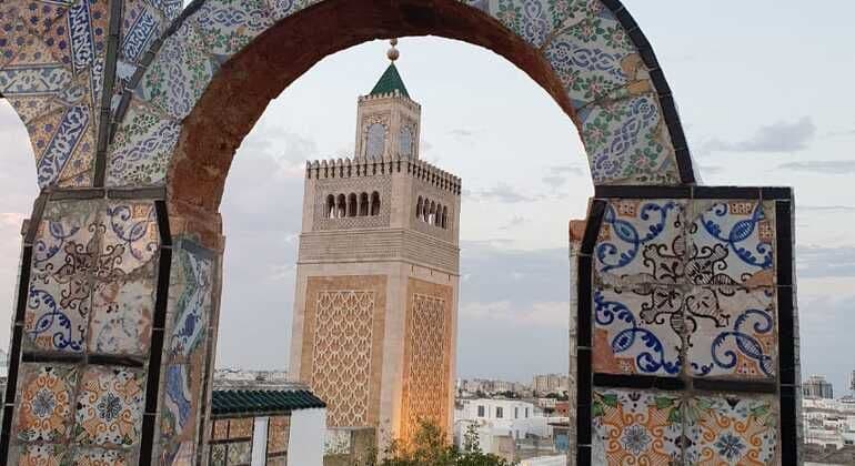 Un minarete de piedra con tejado verde, enmarcado por un arco ornamentado de azulejos coloridos, con vistas a un paisaje urbano al atardecer.