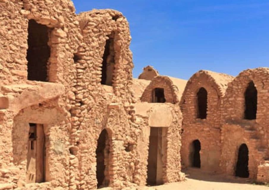 Edificios de piedra desgastados, de color arena, con puertas y ventanas arqueadas, bajo un cielo azul claro.