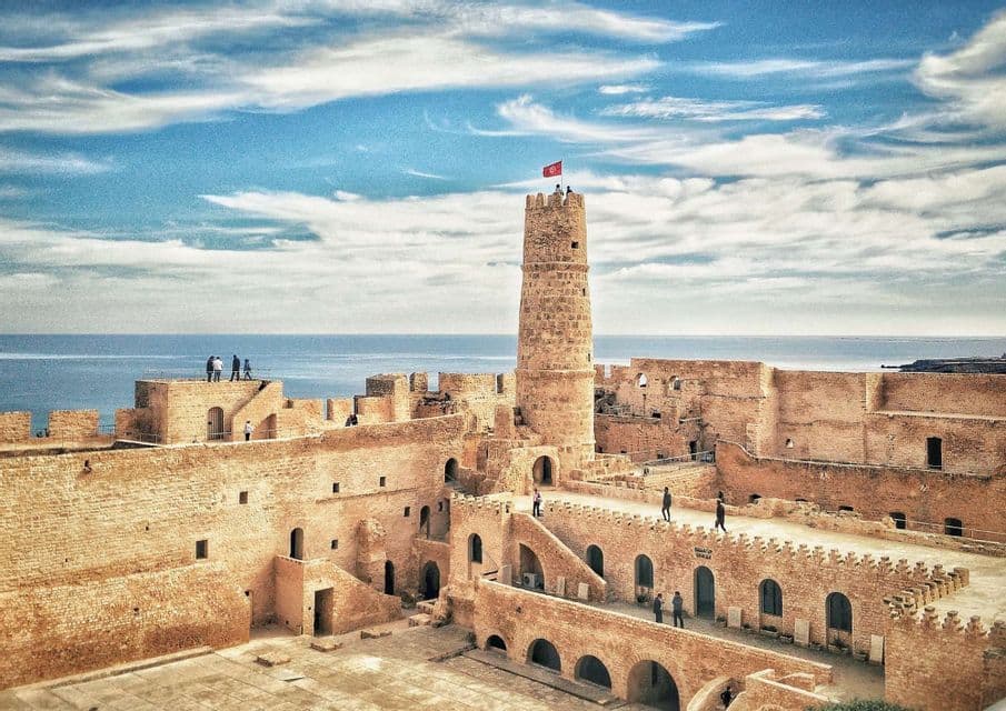Un viaje en grupo de WeRoad explora una fortaleza histórica de piedra con una torre central y una bandera, con vistas al mar.