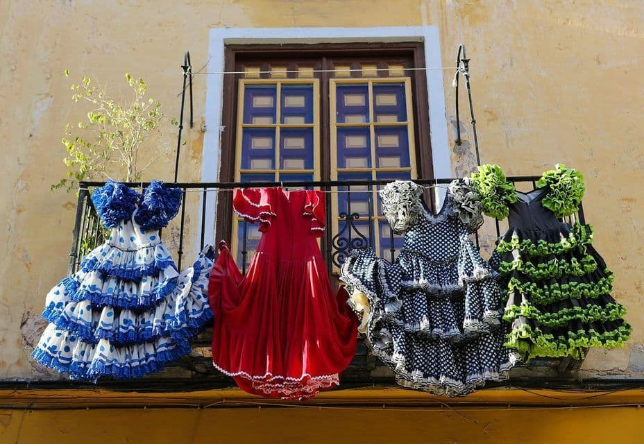 Four colorful, ruffled flamenco dresses hanging on a clothesline on a balcony in front of a window.