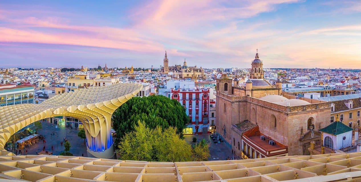 A panoramic view of a city with historic architecture and a large modern wooden structure under a colorful sunset sky.