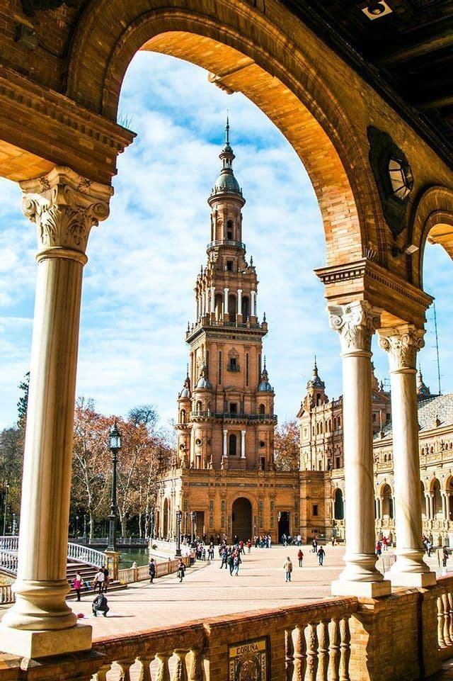 Una torre in mattoni alta e riccamente decorata, vista attraverso un grande arco, con persone che passeggiano nella vasta piazza sottostante sotto un cielo parzialmente nuvoloso.