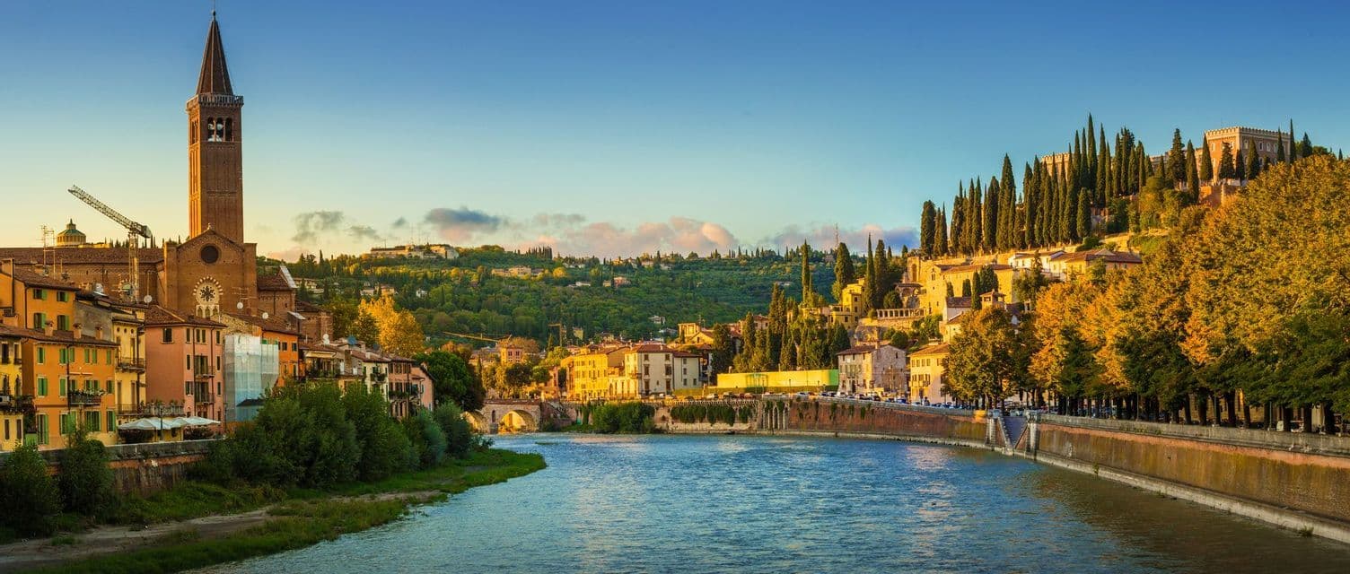 Una vista panoramica di una città con un'alta torre campanaria in mattoni e edifici colorati che fiancheggiano un ampio fiume al tramonto.