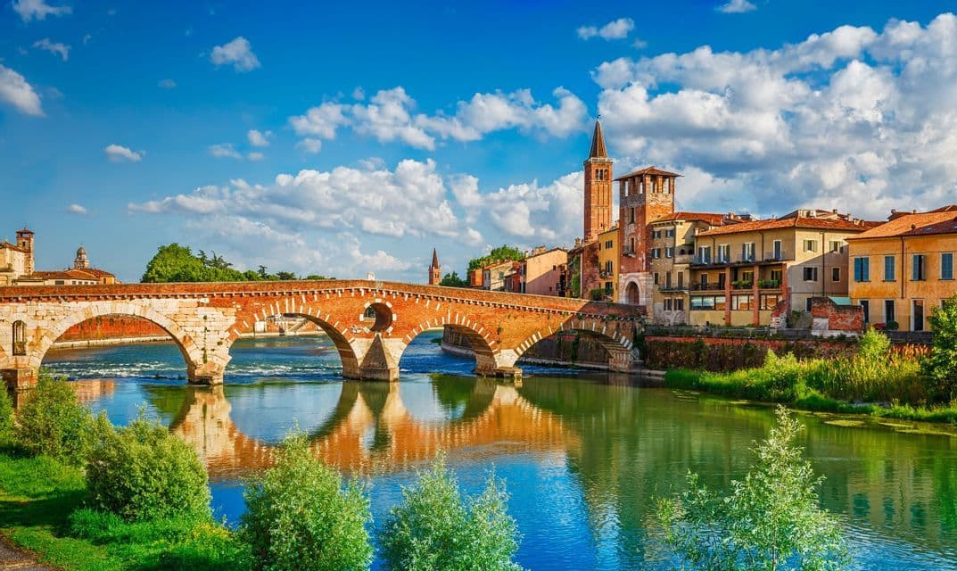 Un ponte ad arco in mattoni attraversa un ampio fiume di fronte a un paesaggio urbano storico con una torre, sotto un cielo blu parzialmente nuvoloso.