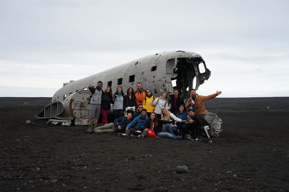 Un groupe WeRoad pose pour une photo devant un avion échoué sur une plage de sable noir sous un ciel nuageux.