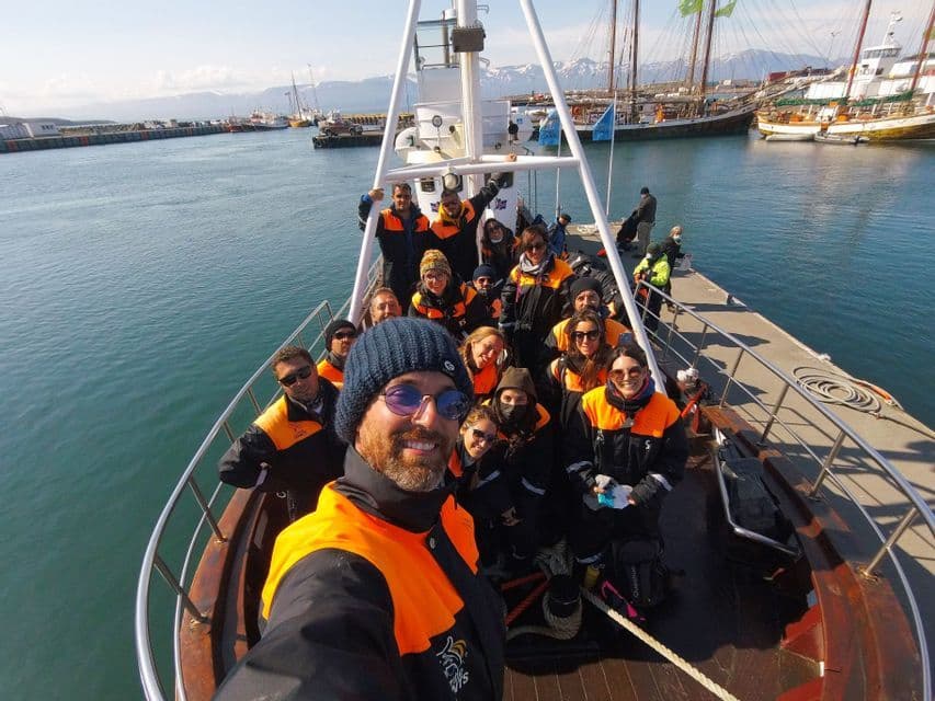 Un homme avec un bonnet prend un selfie avec un groupe WeRoad portant des gilets de sauvetage sur le pont d'un bateau dans un port ensoleillé.