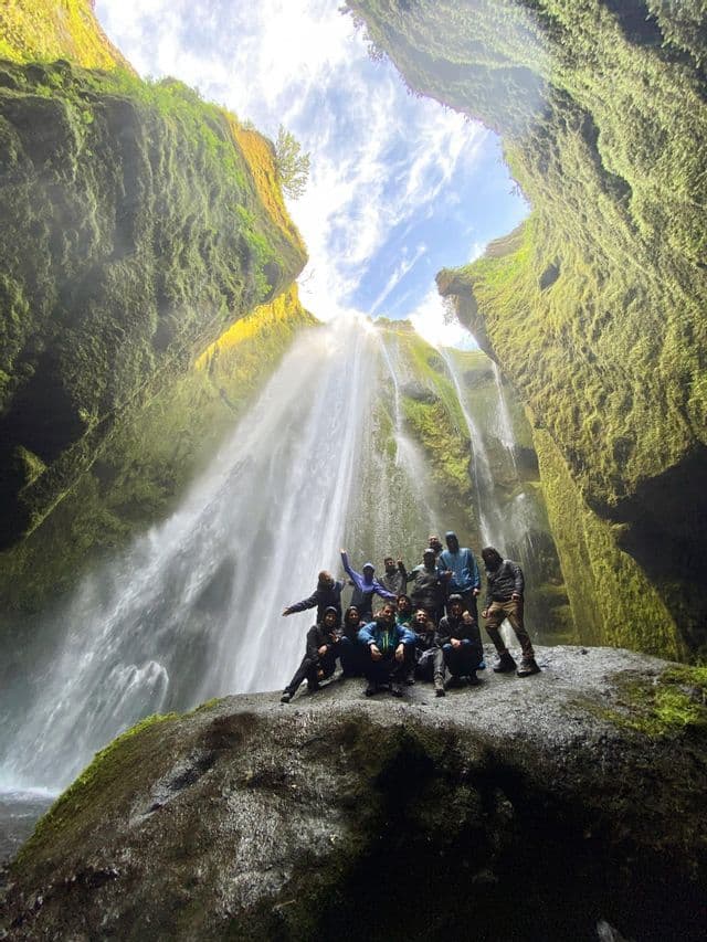Un groupe WeRoad en voyage pose sur un grand rocher devant une puissante cascade à l'intérieur d'un canyon verdoyant et moussu.