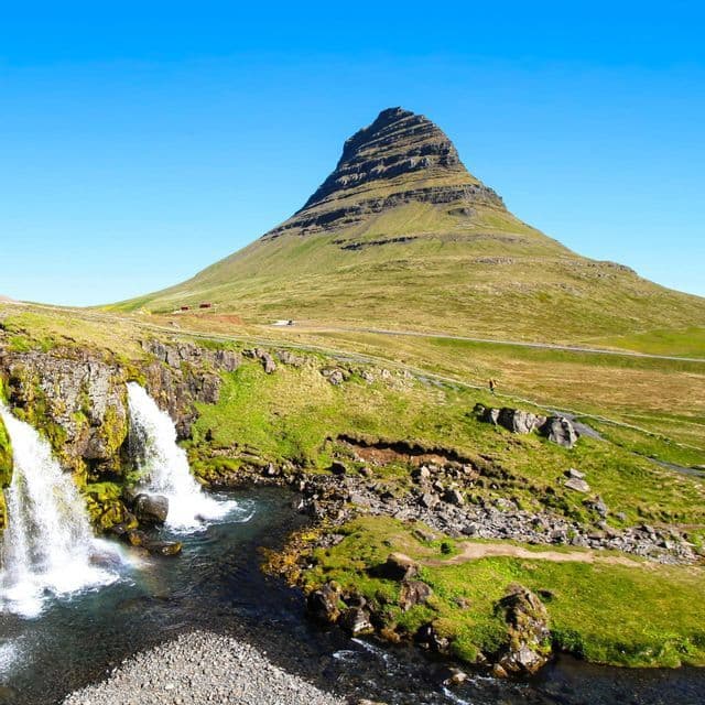 Two waterfalls cascade down a mossy cliff into a stream, with a grassy, cone-shaped mountain behind them under a clear sky.