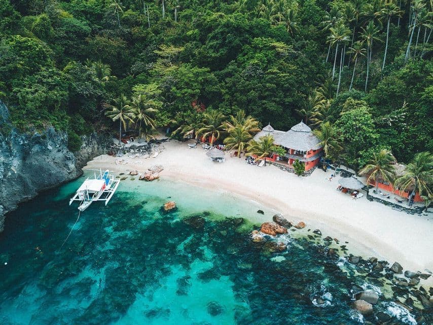 Vue aérienne d'une plage de sable blanc isolée, avec un bâtiment au toit de chaume, bordée d'une forêt dense et d'eaux turquoise limpides avec un catamaran amarré.