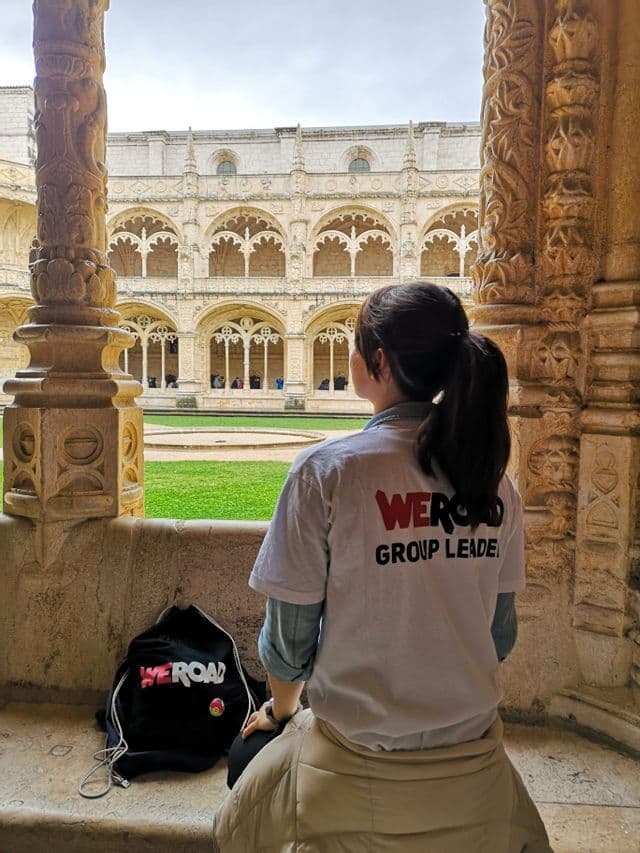 A WeRoad group leader sits on a stone ledge, viewed from behind, looking out at a historic cloister with a grassy courtyard.