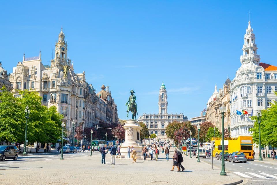 A wide, bustling city square centered around an equestrian statue, flanked by ornate historical buildings under a clear blue sky.