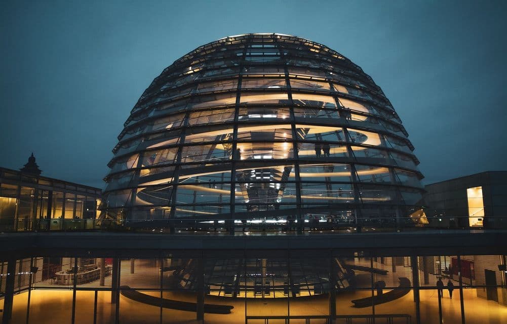 The illuminated glass dome of a modern building at night, with silhouettes of people visible on an internal walkway.