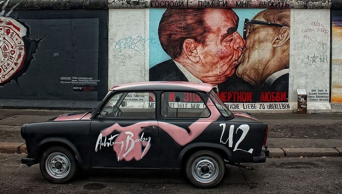 A black car with "U2" and "Achtung Baby" painted on its side, parked in front of a large mural of two men kissing on a concrete wall.