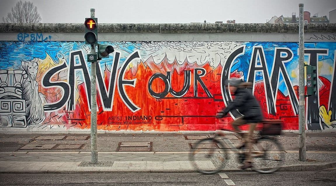 Une personne floue à vélo longe un trottoir devant une fresque murale colorée où l'on peut lire 'SAVE OUR ART'.