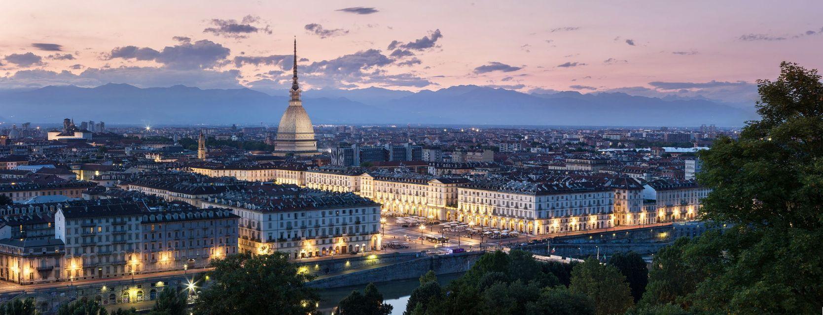 Un vasto paesaggio urbano caratterizzato da una caratteristica guglia a cupola, illuminato al crepuscolo con montagne in lontananza sotto un cielo nuvoloso.