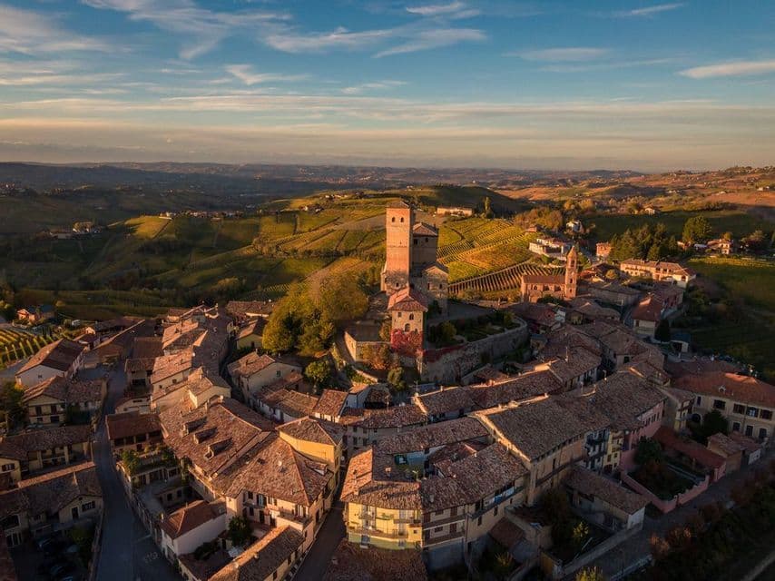 Una vista aerea di un borgo storico con una torre centrale, immerso tra dolci colline coperte di vigneti al tramonto.