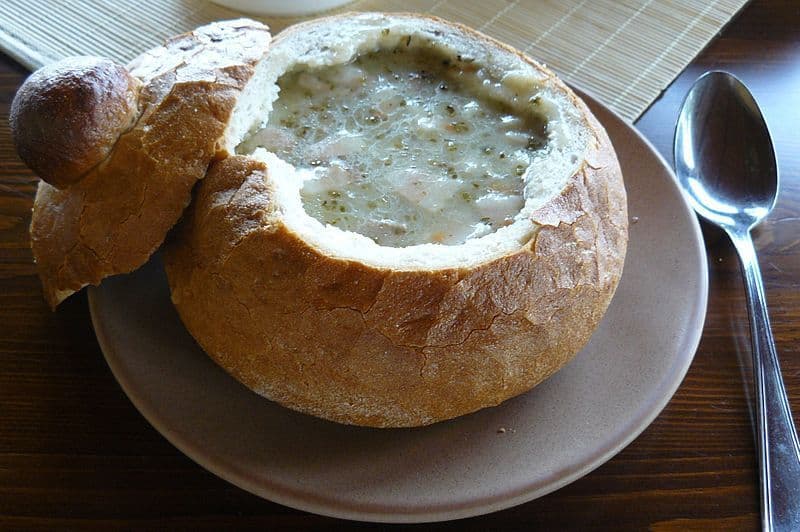 Une soupe onctueuse servie dans un bol de pain creusé avec son couvercle, présentée sur une assiette à côté d'une cuillère.