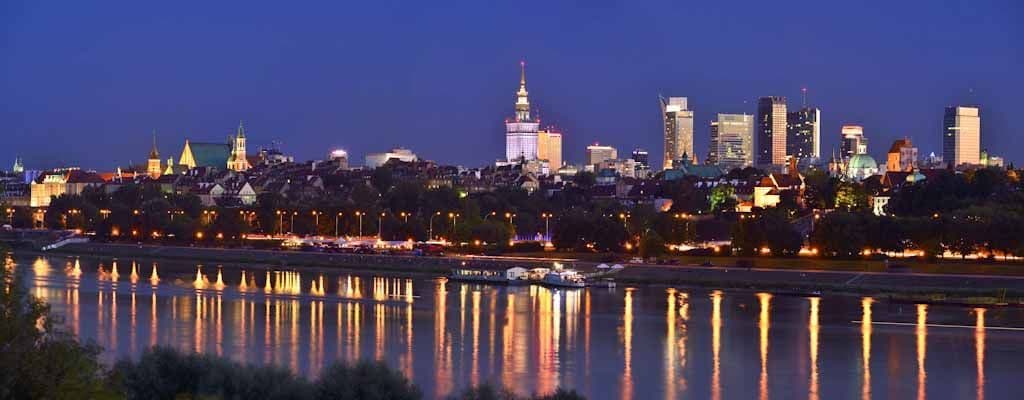 A city skyline illuminated at twilight, viewed from across a river with lights reflecting on the water's surface.
