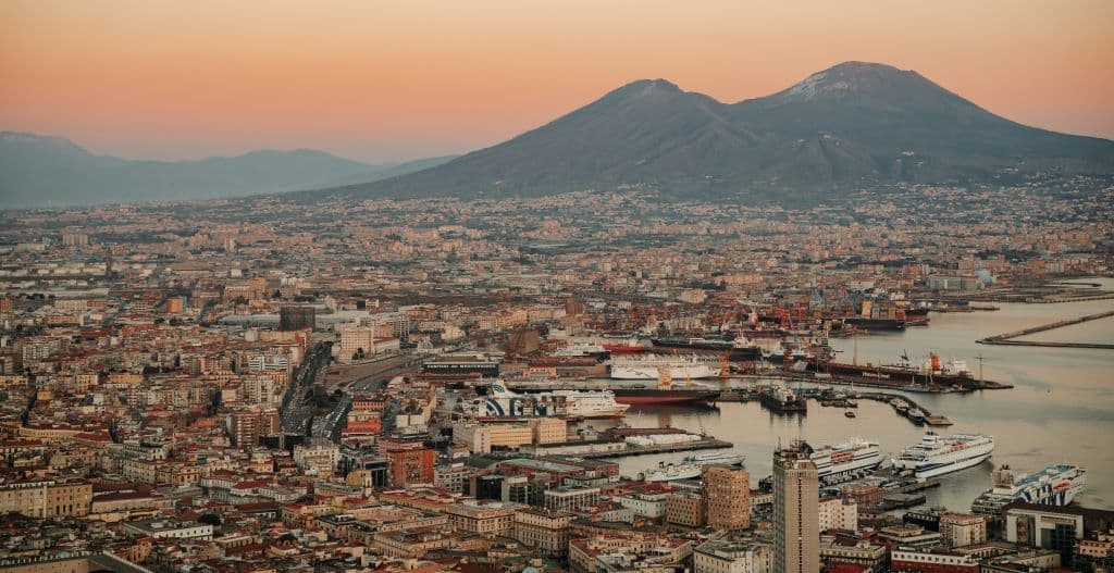 Una vista aérea de una extensa ciudad costera y su concurrido puerto al pie de un gran volcán bajo un cielo de atardecer de tonos cálidos.