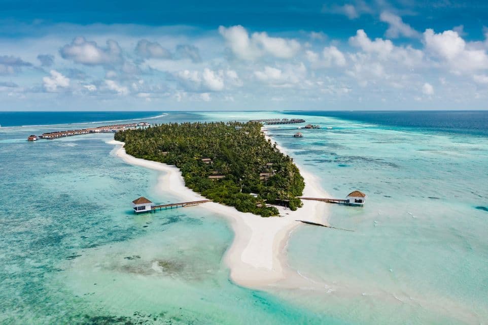 Vue aérienne d'une île tropicale avec des palmiers, des bancs de sable blanc et des bungalows sur pilotis dans un océan turquoise sous un ciel partiellement nuageux.
