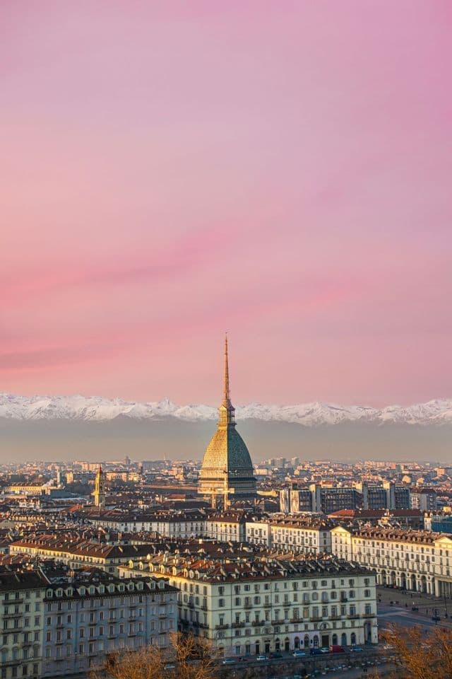Una vista aerea di una città con un edificio centrale a cupola, sullo sfondo di montagne innevate sotto un cielo rosa al tramonto.
