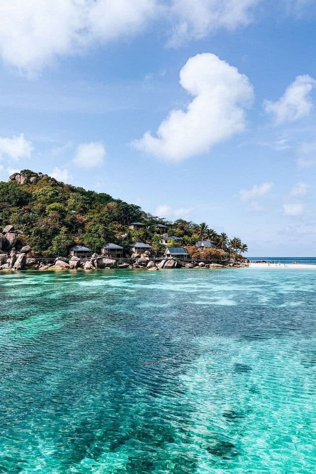 Une île verte et rocheuse avec des bungalows le long du rivage, entourée d'une eau turquoise claire sous un ciel partiellement nuageux.