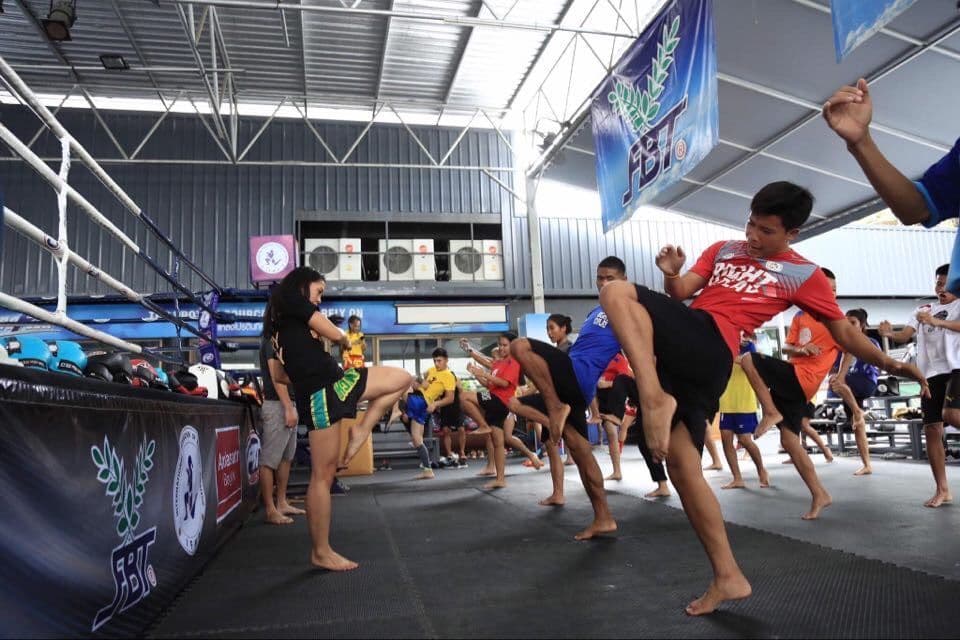 Un voyage de groupe WeRoad avec des participants s'entraînant aux high kicks dans une salle de Muay Thai, à côté d'un ring de boxe.